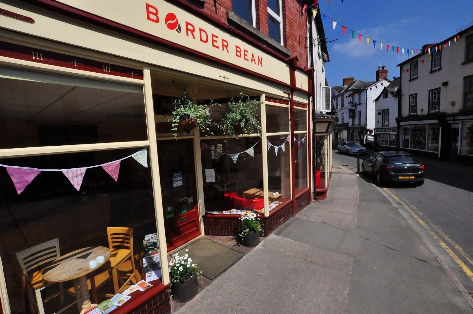 The shopfront seen along the pavement: bunting across the windows, plants and café chairs outside