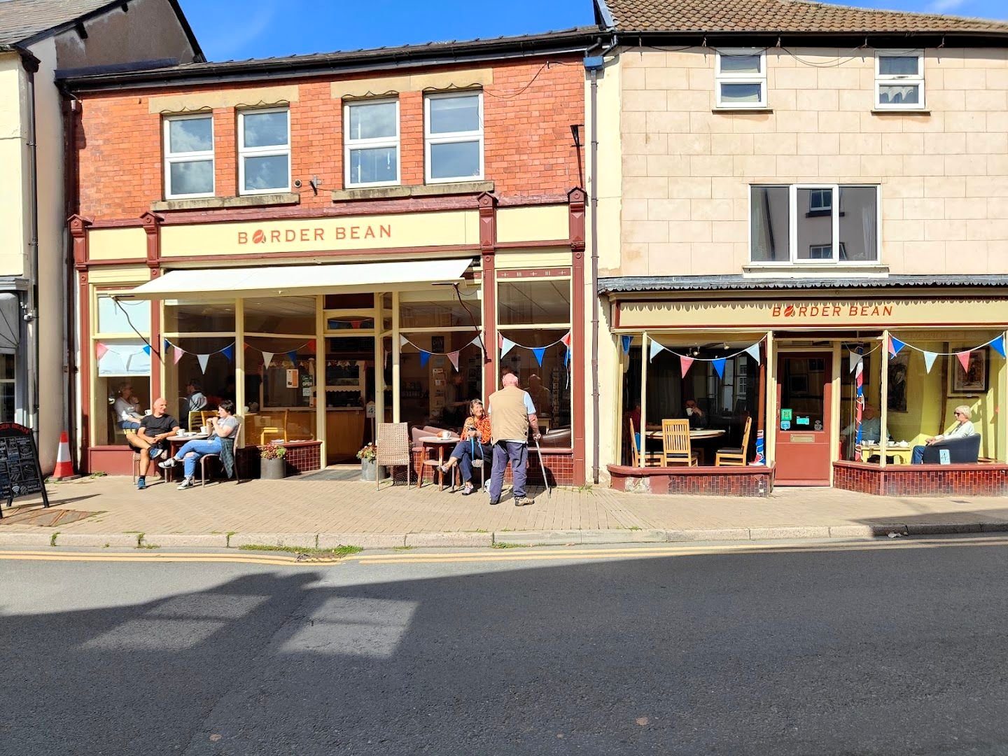 Border Bean from across the High Street: a cream and red fascia over two shop windows, with people at tables outside